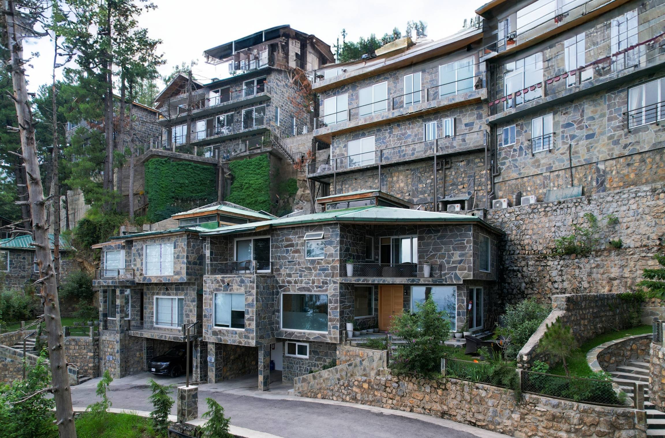Multi-storey stone-clad resort building with balconies and large windows, built into a steep terraced hillside surrounded by pine trees and lush greenery