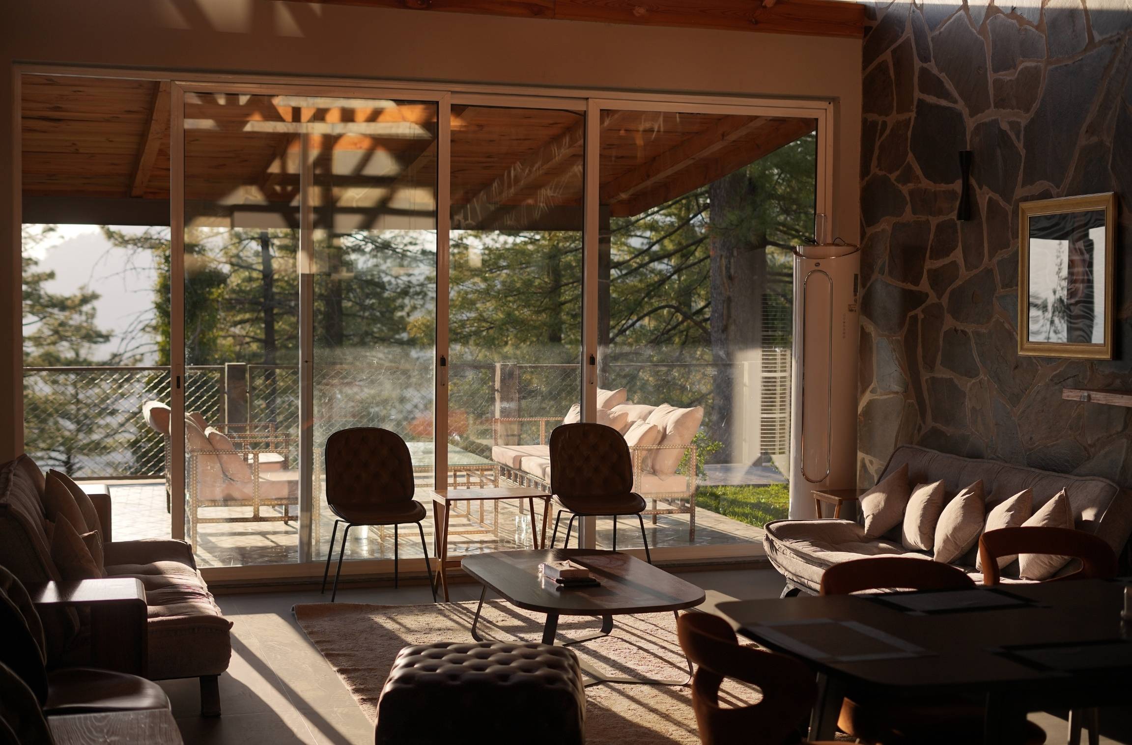 Warm interior living room of a mountain resort with wooden ceiling, stone wall, large glass sliding doors opening to a pine-forested terrace