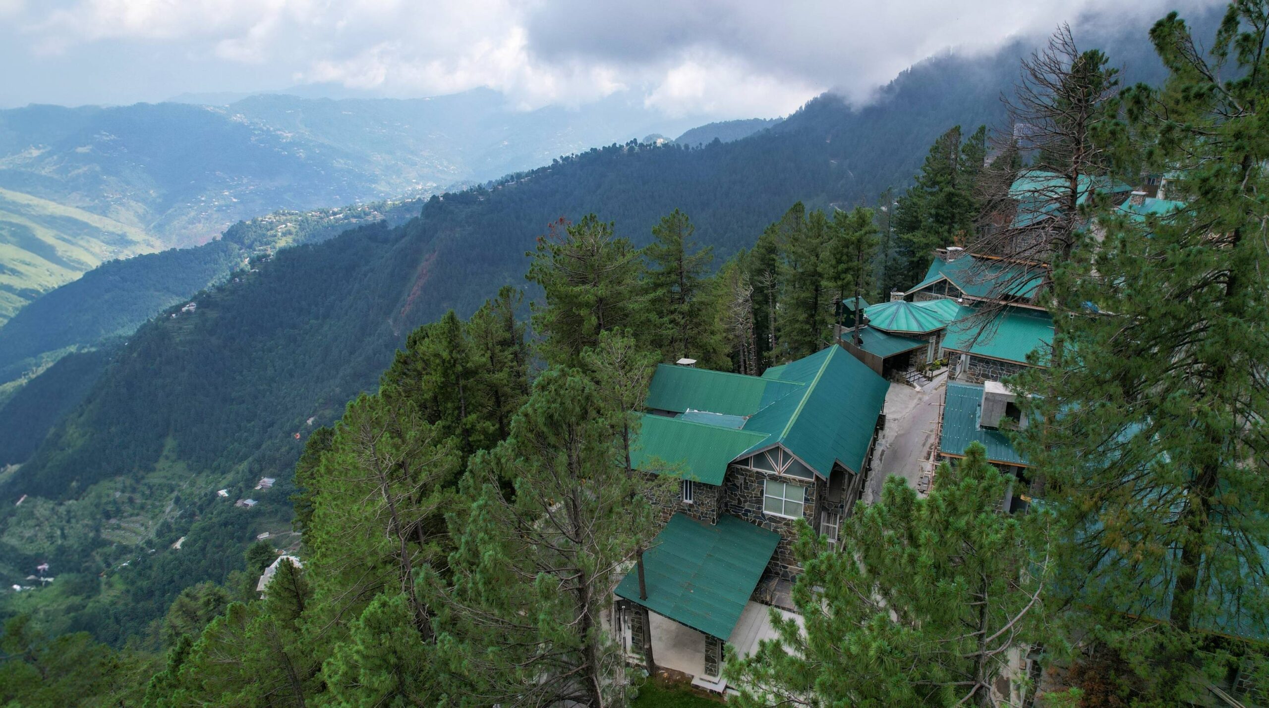 Aerial view of a multi-unit stone resort with green metal rooftops nestled among tall pine trees on a steep mountain slope, with a misty valley below