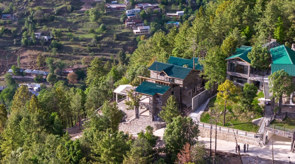 Modern luxury living room with exposed wood beam ceiling, stone fireplace, and panoramic pine forest view in Galiyat villa